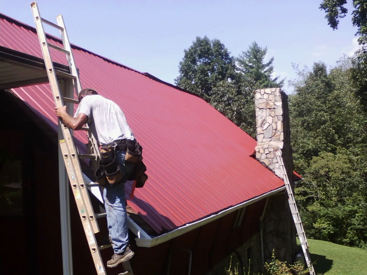 Roofer on a ladder beside a red metal roof and stone chimney — Soffit & Fascia Repair by Roofing Brothers in Shoup